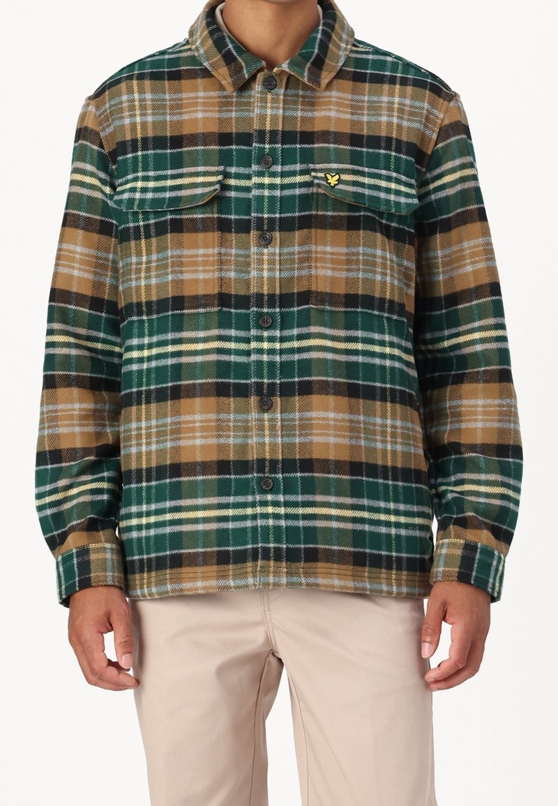 Homme portant une chemise à carreaux verte, marron, noire et blanche avec poches poitrine et pantalon beige, visible du cou aux hanches.