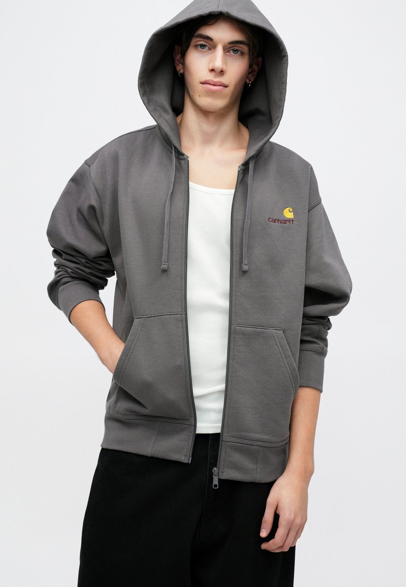 Junge Person, die einen grauen Reißverschluss-Hoodie von Carhartt über einem weißen Tanktop und schwarzen Hosen trägt, steht vor einem schlichten hellen Hintergrund.