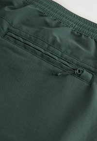 Pantaloni scurți cargo de culoare verde închis, dintr-un material moale, cu un tiv elastic și un buzunar cu fermoar în spate, dotat cu un șiret de strângere.