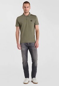 Olijfgroen poloshirt met een kraag en drie knopen, met een cirkelvormig logo. Gecombineerd met grijze jeans en witte sneakers.