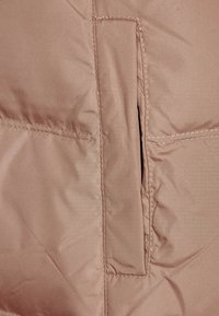 Chaqueta acolchada de suave tela beige con un bolsillo lateral oculto, que presenta un detalle de costuras y una textura lisa.