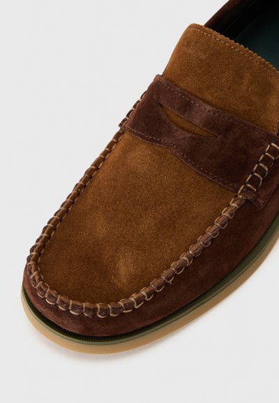 Mocassin en velours brun avec un accent contrastant marron foncé, des détails en couture et une semelle en caoutchouc. Texture lisse et forme arrondie.