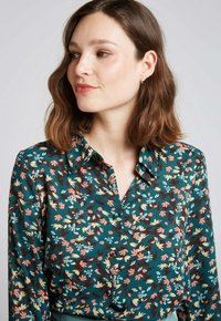 Blouse florale verte à manches longues, col boutonné, ornée d'un motif multicolore de fleurs et de feuilles sur un tissu léger.
