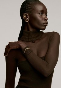 Top de cuello alto acanalado en marrón oscuro con mangas transparentes; acentos de joyería dorada y cabello trenzado; la textura de la piel suave contrasta con el tejido.