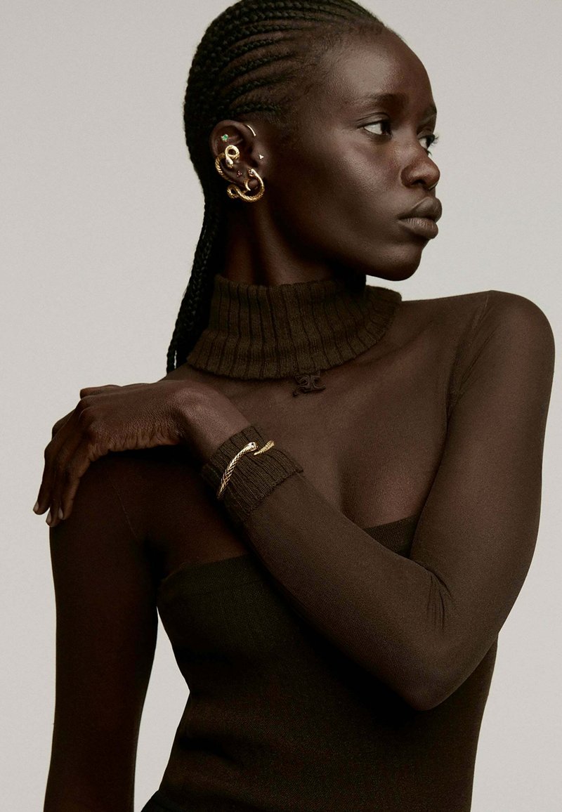 Top de cuello alto acanalado en marrón oscuro con mangas transparentes; acentos de joyería dorada y cabello trenzado; la textura de la piel suave contrasta con el tejido.