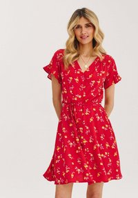 Czerwona sukienka w kwiaty z krótkimi falbaniastymi rękawami, dekoltem w serek, guzikami z przodu i wiązaniem w talii. Posiada wzory z żółtymi i różowymi kwiatami.
