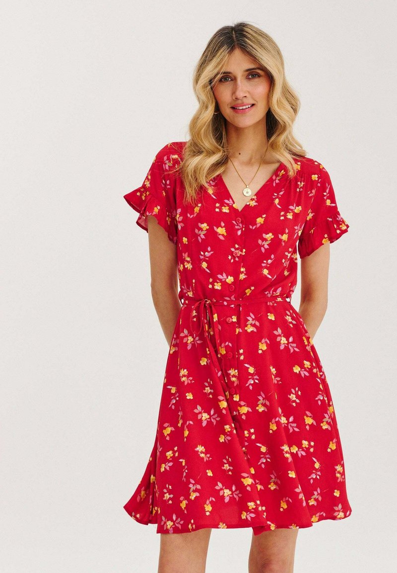 Czerwona sukienka w kwiaty z krótkimi falbaniastymi rękawami, dekoltem w serek, guzikami z przodu i wiązaniem w talii. Posiada wzory z żółtymi i różowymi kwiatami.