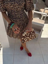 Luipaardprintjurk, bruine gevlochten handtas met grote beige kralen, rode ballerina's, en meerdere ringen en armbanden om de pols. Achtergrond van witte tegels.