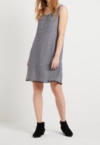 Gestreiftes ärmelloses Kleid aus marineblauem und weißem Stoff, mit gerafftem Ausschnitt und schwarzem Spitzenbesatz am Saum. Getragen mit schwarzen Knöchelstiefeln.