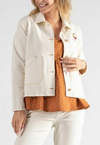 Weiße Jeansjacke mit goldenen Knöpfen und gesticktem Blumenmotiv. Trägt man über einer rostfarbenen gemusterten Bluse mit ausgestelltem Saum.