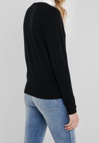 Donna che indossa un maglione nero a maniche lunghe e jeans azzurri chiari, in piedi contro uno sfondo grigio chiaro uniforme.