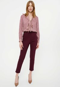 Patroon blouse met een striknek, uitgevoerd in een bordeauxrood en ivoorkleurig kleurenschema, gecombineerd met tapse bordeauxrode broeken en beige hakken.