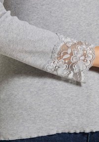 Grijze geribbelde stoffen shirt met delicate bloemenkant accenten bij de manchetten, dat een gemêleerd patroon en een iets rafelige zoom toont.
