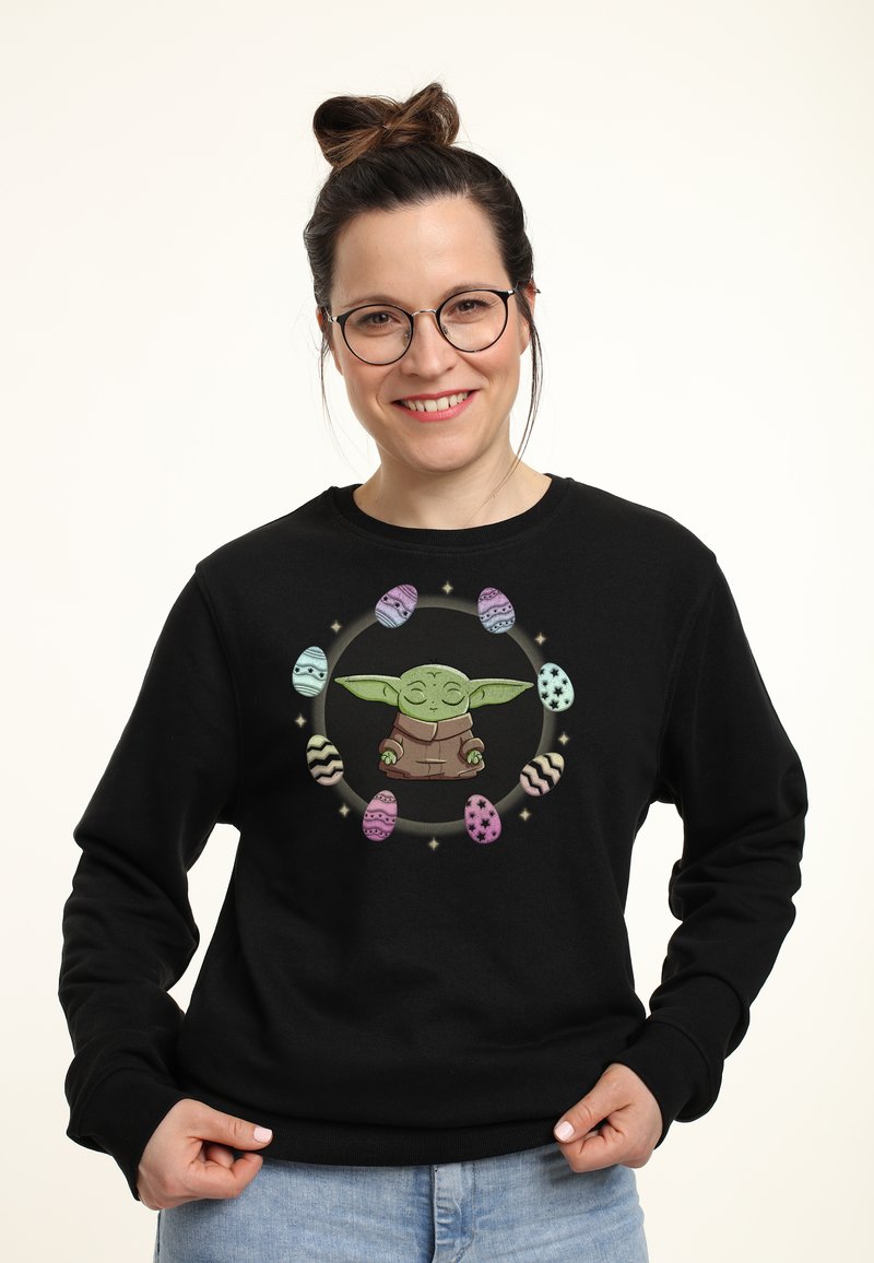 Sudadera negra con un gráfico central de un personaje verde con orejas grandes, rodeado de coloridos huevos de Pascua en varios patrones.