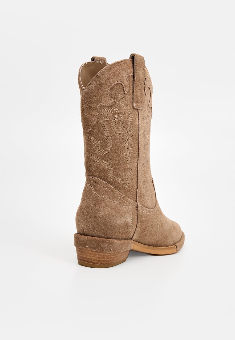 Bota de cowboy de ante marrón con detalles bordados, puntera redondeada y un tacón bajo de madera. Lengüetas laterales para facilitar su uso.