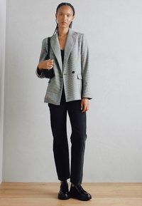 Grau gestreifter Blazer mit doppelter Knopfleiste, schwarzen Akzenten, kombiniert mit schwarzen Slim-Fit-Hosen und schwarzen Schnürschuhen.