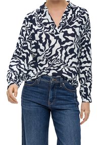 Person med långärmad blus i svart och vitt abstrakt mönster som är instoppad i högmidjade blå jeans, händerna avslappnat vid sidorna.