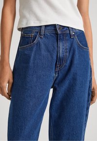 Vaqueros de denim azul de tiro alto y corte holgado, con cinco bolsillos, costuras naranjas contrastantes y un cierre de botón de metal en la cintura.