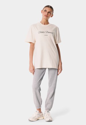 Kobieta w oversize kremowym T-shircie z napisem "Pilates Princess", szarych spodniach dresowych i białych trampkach, stojąca na białym tle.