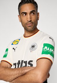 Balts sporta krekls ar melnām detaļām, sarkanu un dzeltenu apmali, uz tā attēlotas Puma, Lidl un Alsco logotipi. Redzams arī Vācijas valsts ģerbonis.