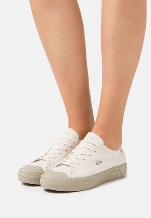 Beige canvas sneakers with a textured rubber sole, featuring a lace-up design and a small embroidered logo on the side.
