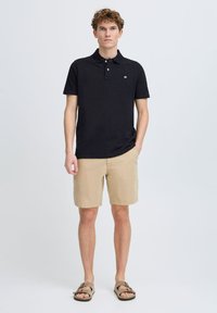 Chemise polo noire avec un col, manches courtes et un petit logo ; assortie à un short beige et des sandales marron. Style simple et décontracté.