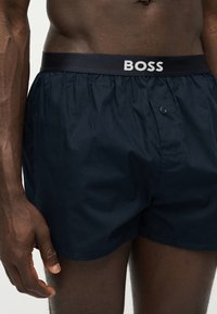 Gros plan d'une personne portant un boxer bleu foncé avec une ceinture noire comportant le mot « BOSS » en lettres blanches.
