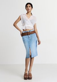 Femme portant un top transparent blanc, une jupe en denim bleu clair jusqu'au genou avec une fente à l'avant et une large ceinture tissée marron, ainsi que des chaussures à plateforme marron.