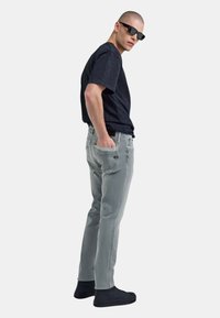 Camiseta corta azul oscuro, jeans de mezclilla grises, zapatillas negras y gafas de sol. El modelo está de perfil con las manos en los bolsillos.