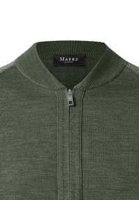 Zbliżenie na oliwkowy sweter na zamek z prążkowanym kołnierzem i czarną metką "Maerz Muenchen" wewnątrz dekoltu.