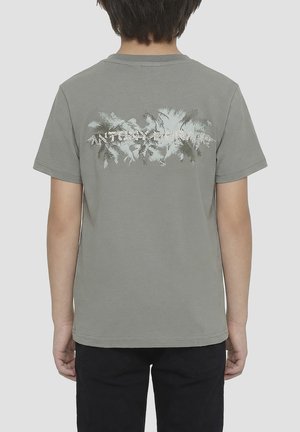 Enfant portant un t-shirt gris avec un motif de palmier délavé et un texte sur le haut du dos, associé à un pantalon noir, debout de dos.