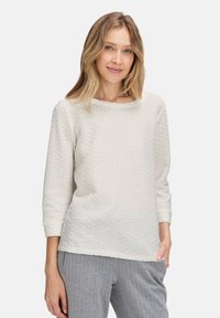 Femme aux cheveux blonds portant un haut à manches longues texturé blanc et un pantalon à rayures grises, se tenant debout avec une main dans la poche.