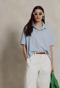 Šviesiai mėlynas polo marškinėliai su apykakle, balti stačiakampiai saulės akiniai, beige spalvos diržas, balti kelnės ir žalia rankinė laikoma vienoje rankoje.