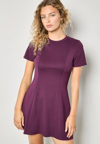 Kurzes Kleid mit kurzen Ärmeln in tiefem Lila, aus elastischem Stoff. Hervorgehoben durch ein tailliertes Oberteil mit sauberen Nähten und einem ausgestellten Rock, mit glatter Textur.