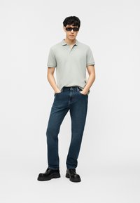 Άνδρας που φοράει ανοιχτό γκρι πόλο πουκάμισο, σκούρο μπλε τζιν, μαύρα chunky μποτάκια και μαύρα ορθογώνια γυαλιά ηλίου, στέκεται με τα χέρια στις τσέπες.