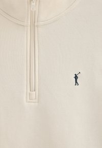Suéter de media cremallera color Beige claro, hecho de una tela suave, que presenta un sutil patrón texturizado y un pequeño logotipo oscuro de un golfista en el lado izquierdo del pecho.