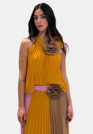 Donna con capelli lunghi e scuri che indossa un vestito a pieghe giallo senape con dettagli floreali marroni e una cintura rosa, in piedi davanti a uno sfondo semplice.