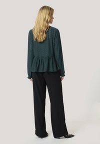 Dunkelgrünes Peplum-Top mit kompliziertem Muster, langen Ärmeln und elastischen Bündchen, kombiniert mit weit geschnittenen schwarzen Hosen und Absatzschuhen.