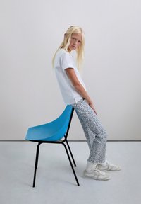 Tricou alb cu mânecă scurtă, pantaloni albstri cu model floral și adidași albi purtați de o fată care se sprijină de un scaun albastru într-un mediu simplu.