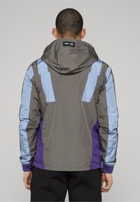 Kapuzensweatshirt mit grauer Basis, versehen mit blauen und violetten Akzenten. Strukturiertes Finish, sichtbare Nähte und verstellbare Details an den Bündchen.