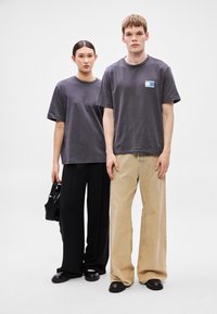 Dois modelos vestem t-shirts de manga curta e oversize de cor cinza escura. A mulher segura uma pequena bolsa preta, combinada com calças largas pretas. O homem usa calças bege claro.