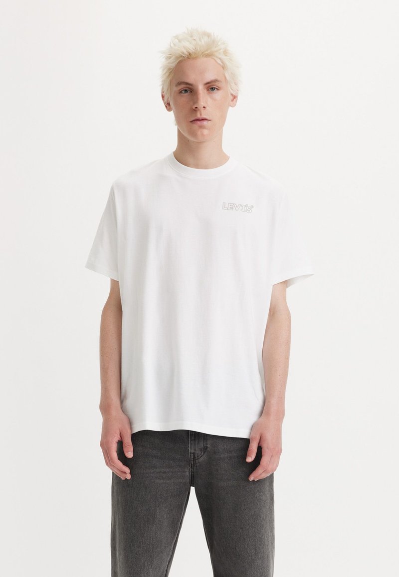 Camiseta de algodón blanca con cuello redondo, que presenta un pequeño logo en gris "LEVI'S" en el lado izquierdo del pecho, combinada con jeans gris oscuro.