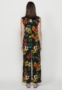 Jumpsuit floreale con sfondo nero; presenta vivaci fiori rossi e bianchi, foglie verdi, maniche a cappuccio e una schiena a chiave. Design a gamba larga.