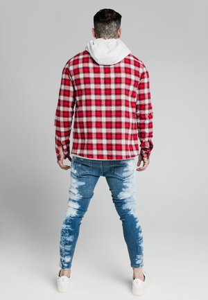 Mężczyzna z czarnym tatuażem kwiatowym na szyi, ubrany w czerwoną koszulę w kratę na białej bluzie z kapturem, jasne jeansy typu skinny z efektem wybielania i białe trampki, zwrócony plecami.