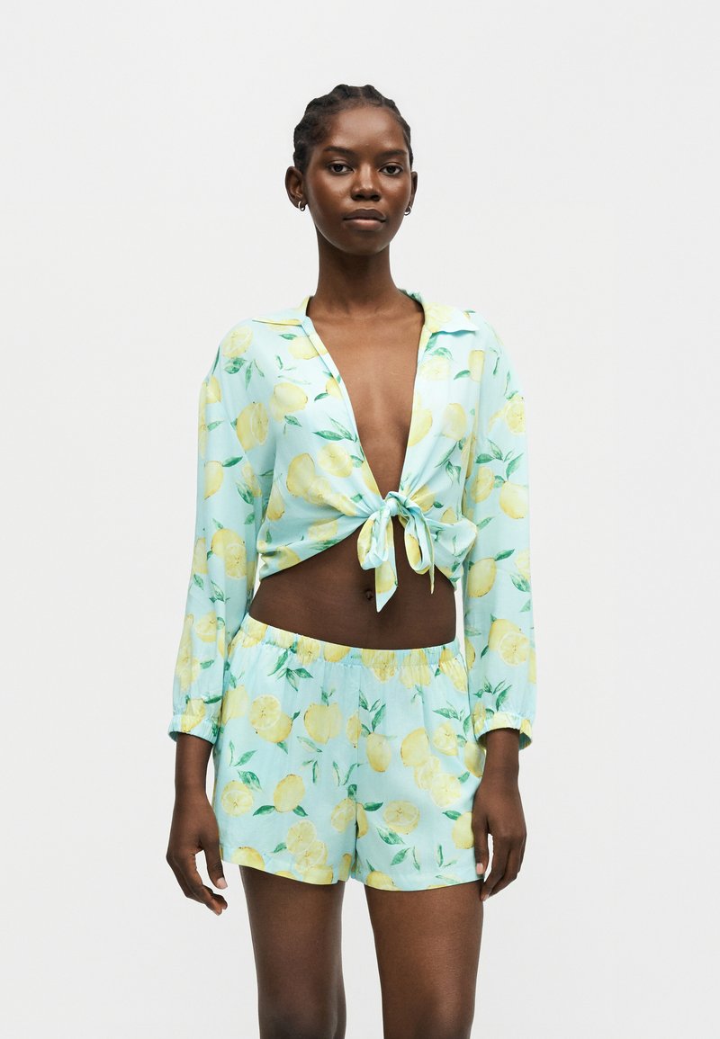 Femme aux cheveux tressés portant une tenue deux pièces bleu clair à imprimé citron jaune, chemise à manches longues nouée et short à taille élastique sur fond blanc.
