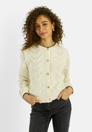 Mujer con cabello rizado que lleva un chaleco de punto color crema abotonado sobre una blusa de manga larga color crema con bordados y jeans negros, con las manos en los bolsillos.