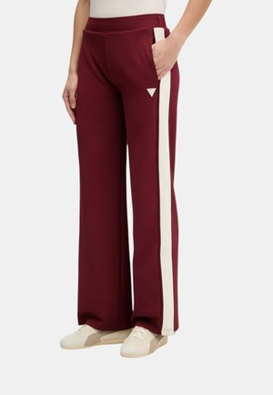 Persona che indossa pantaloni bordeaux a gamba larga con strisce laterali bianche e sneakers bianche, mano nella tasca destra, in piedi contro uno sfondo bianco.