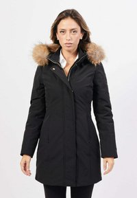 Donna che indossa un cappotto invernale nero di lunghezza media con cappuccio bordato di pelliccia, in piedi su uno sfondo bianco semplice.