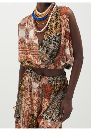 Personne à la peau foncée portant un crop top sans manches multi-motifs et un pantalon dans des tons automnaux, accessoirisée avec des colliers perlés colorés superposés.