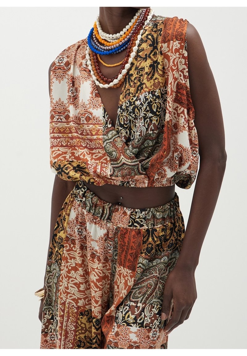 Personne à la peau foncée portant un crop top sans manches multi-motifs et un pantalon dans des tons automnaux, accessoirisée avec des colliers perlés colorés superposés.